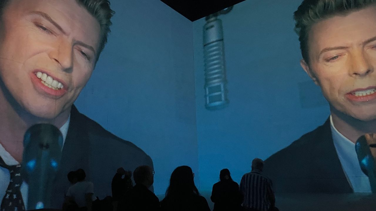 Two walls with projected images of a suited David Bowie singing into a microphone for the exhibition David Bowie: You'e Not Alone at Lightroom, London.