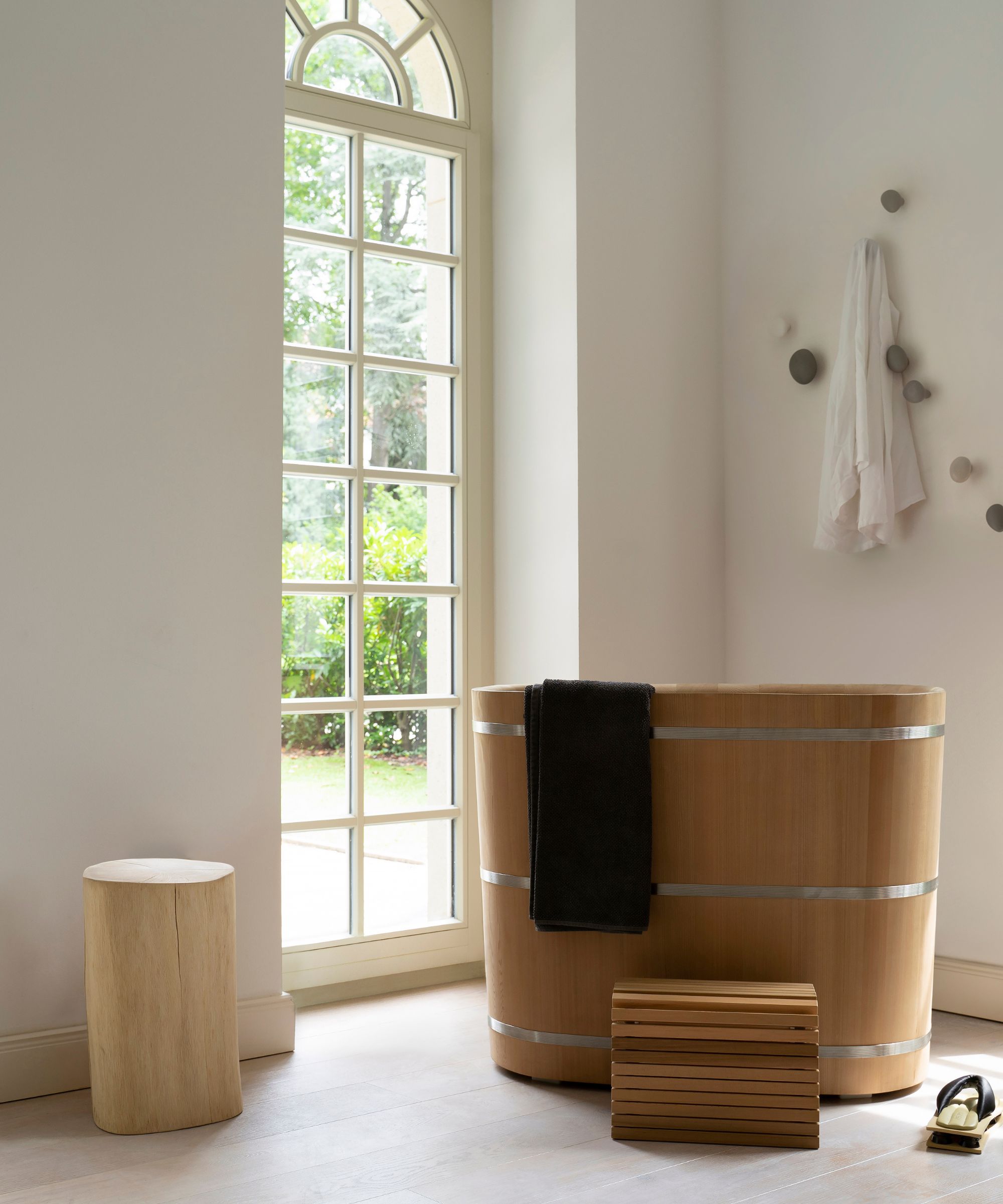 A minimalist, Japanese-inspired bathroom with white walls and light oak floorboards, featuring a deep cylindrical cedar wood ofuro soaking tub with steel banding and a black towel draped over the side. A tall wood stump side table, slatted bath step and round pebble wall hooks holding a white robe sit beside a tall arched Georgian window.
