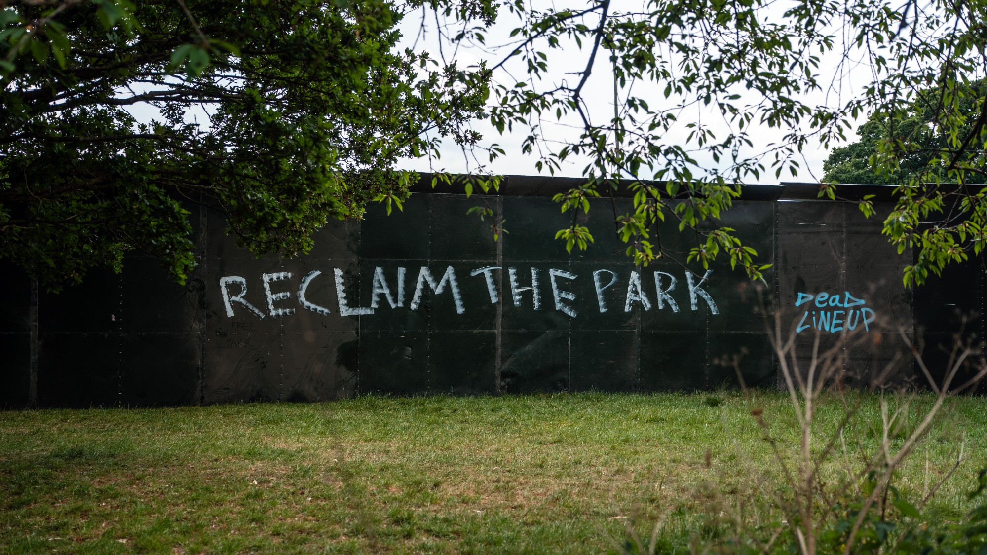 Graffiti reading "reclaim the park" on a metal fence surrounding a temporary festival area in Brockwell Park, London