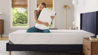 A woman sits on a mattress on a bed frame in a bedroom, holding a Tempur-Pedic Tempur-Cloud Pillow