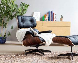 an eames black lounge chair and foot stool in living area with a wooden cabinet, beige rug, a plant and a white throw