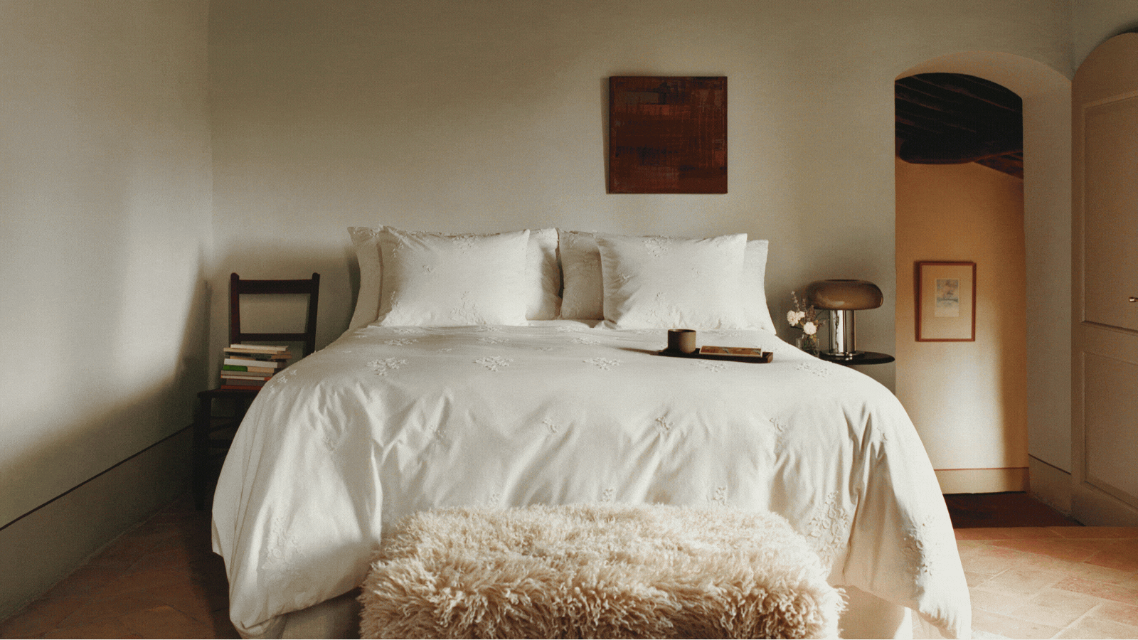 Rotating GIF of four different images that showcase H&M Home's new collection. There is a cozy, white bedroom, a close-up of a table with eyelet linens, a close-up of pink and burgundy striped towels, and a wooden sole table in a neutral hallway.