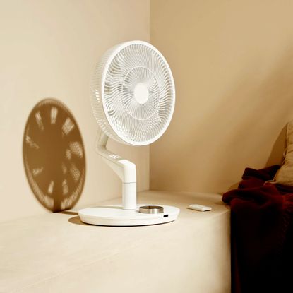 The white Duux Whisper Flex Ultimate Fan sat on a shelf in sunlight that's casting a shadow behind
