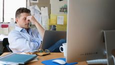 Frustrated male worker sitting at a desk with fingers placed on bridge of nose while in an open plan office space.