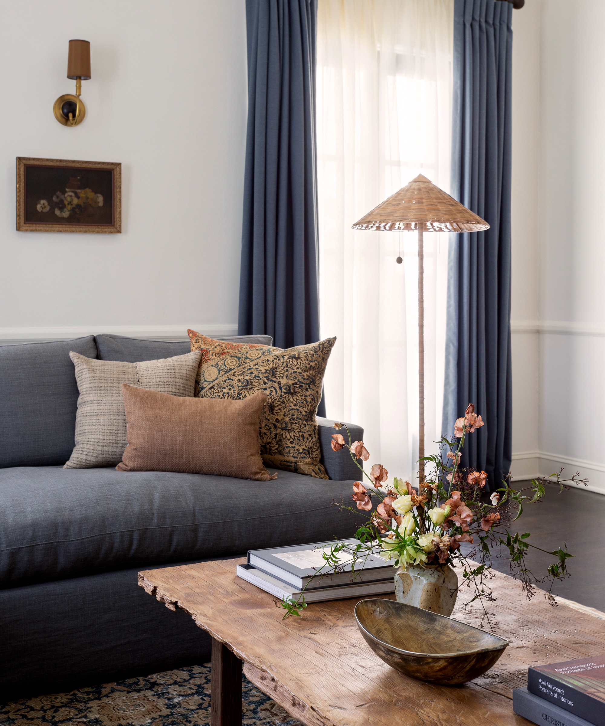 A small living room with white walls, blue drapes, a blue sofa, rattan lamp and a wooden coffee table with fresh flowers on top.