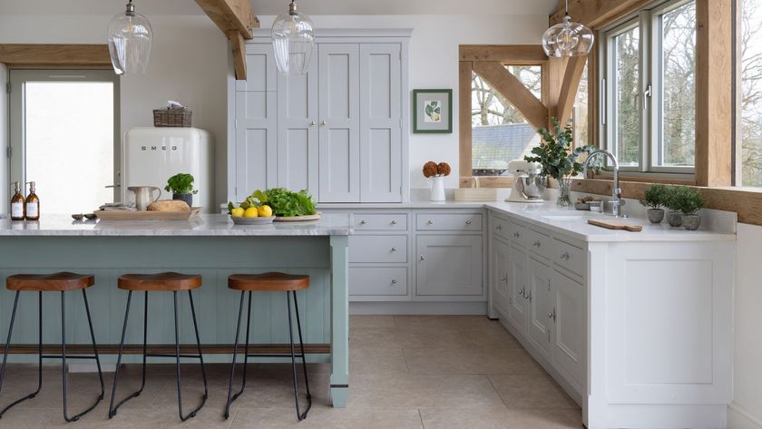 A bright farmhouse-style kitchen with pale grey cabinets, a white worktop and exposed wooden beams. A soft green island with wooden stools sits centrally. Glass pendant lights, fresh greenery and large windows create an airy, welcoming cooking space.