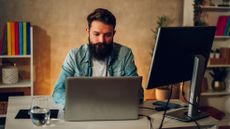 A bearded man sitting at a table that has a laptop, second monitor and a phone