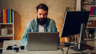 A bearded man sitting at a table that has a laptop, second monitor and a phone