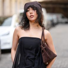 Xiayan Guo wears a brown suede beret with medium-length curly black hair and a middle part, styled with a strapless black jumpsuit featuring a sheer draped overlay. A dark brown suede shoulder bag and silver bangles complete the visible accessories. This is a mid shot, during a street style fashion photo session, on May 07, 2025 in Paris, France. (Photo by Edward Berthelot/Getty Images)