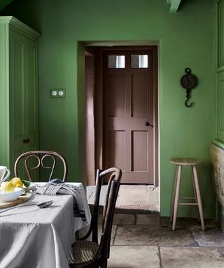 Traditional kitchen-dining space with green painted walls, stone flooring, dining table with white tablecloth, dark wooden dining chairs with rattan seats, light wooden bar stool, green pendant, looking through to hallway with brown front door