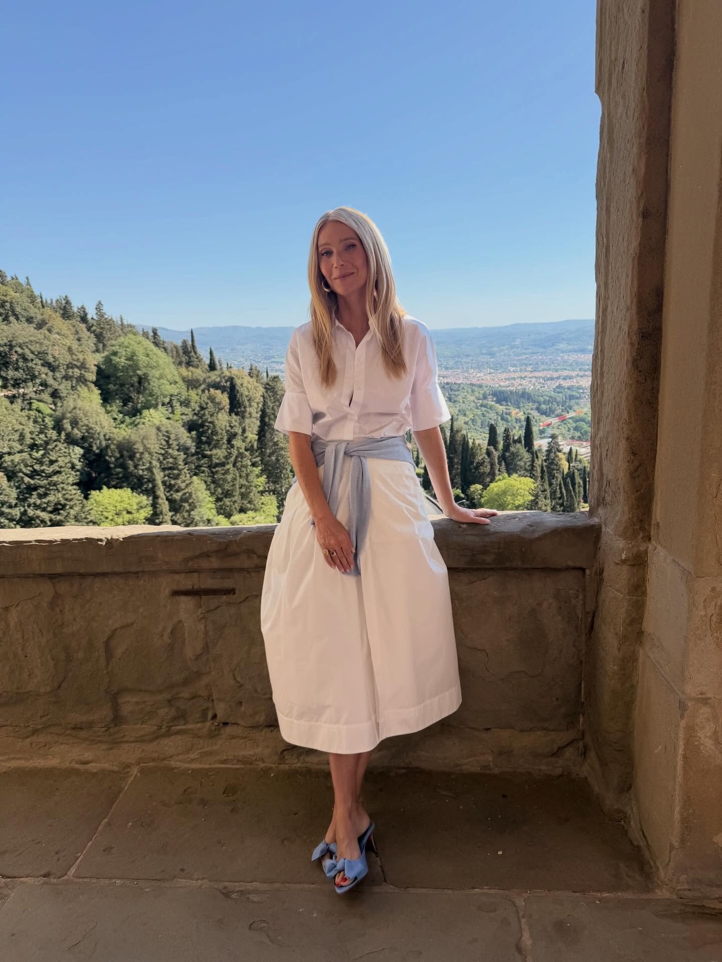 Gwyneth Paltrow poses for a photo outside wearing a white cotton button-down dress with a sky blue jumper wrapped around her waist and a pair of sky blue heels.