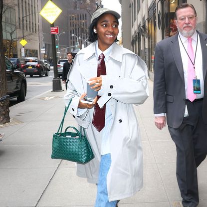 Ayo Edebiri wearing a grey trench coat with a leather neck tie, blue jeans, a green handbag, and a baseball hat