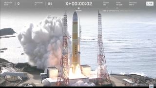 a yellow and white rocket lifts off from a seaside launch pad during the day.