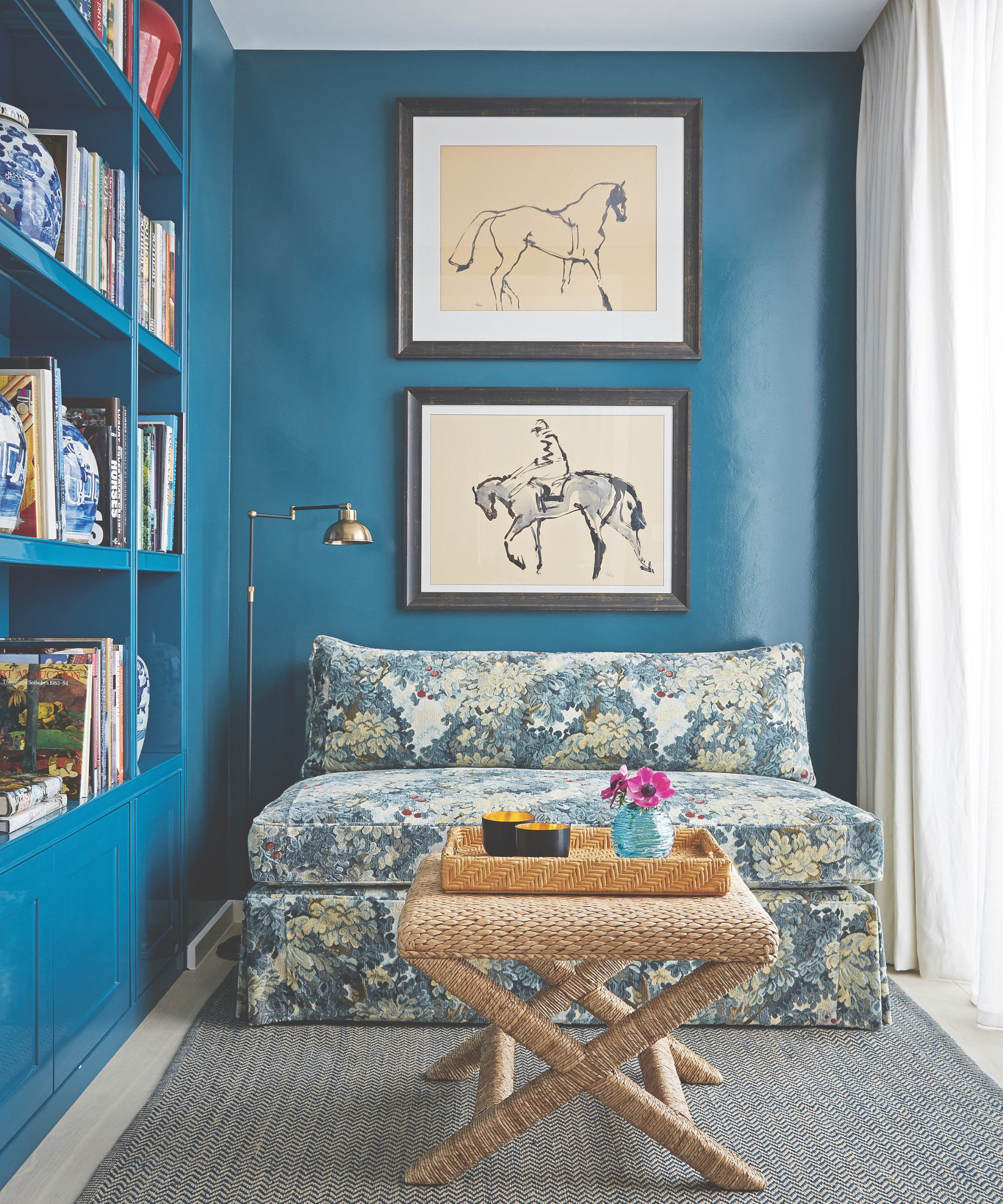 Blue snug room with equestrian prints on wall, floral loveseat, rattan coffee table and built-in bookshelves