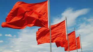 Four red flags on silver poles against a blue sky with light fluffy clouds