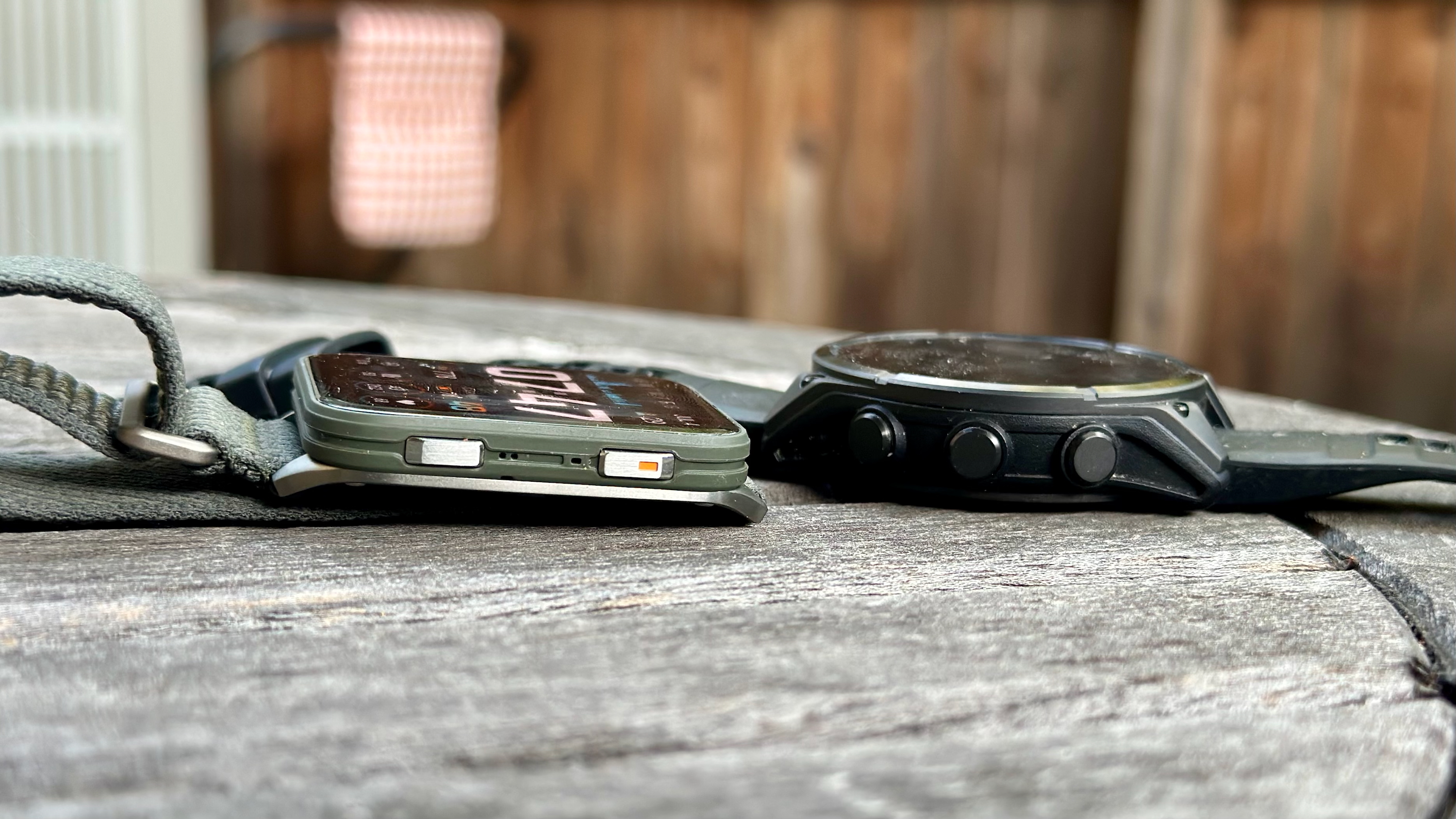 The Garmin Venu X1 (left) and Fenix 8 (right) sitting aside each other on a table, showing the difference in design and thickness.
