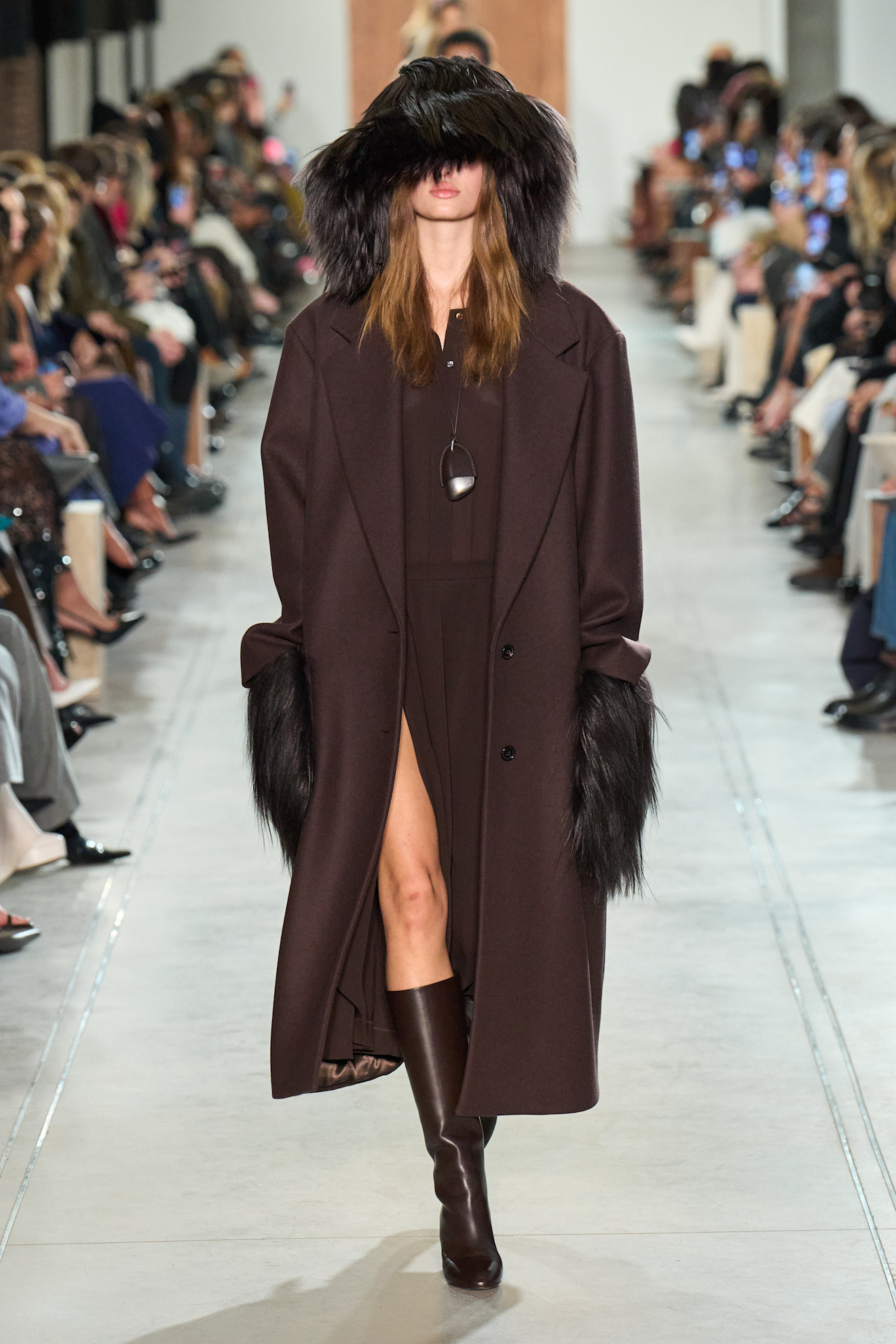 Fur accessory trend shown in Michael Kors' fall-winter 2025 collection in a photo of a model walking down the runway wearing a dark brown fur hat with a chocolate brown coat layered over a matching shirt dress styled with matching knee-high boots and matching furry gloves