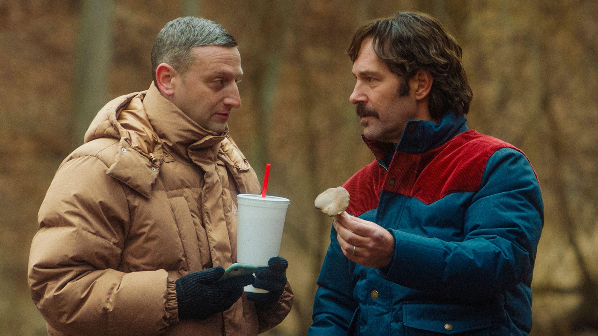 Tim Robinson (left) in brown puffa jacket, holding a fast food drink, Paul Rudd (right) in a blue and red puffa jacket holding a mushroom