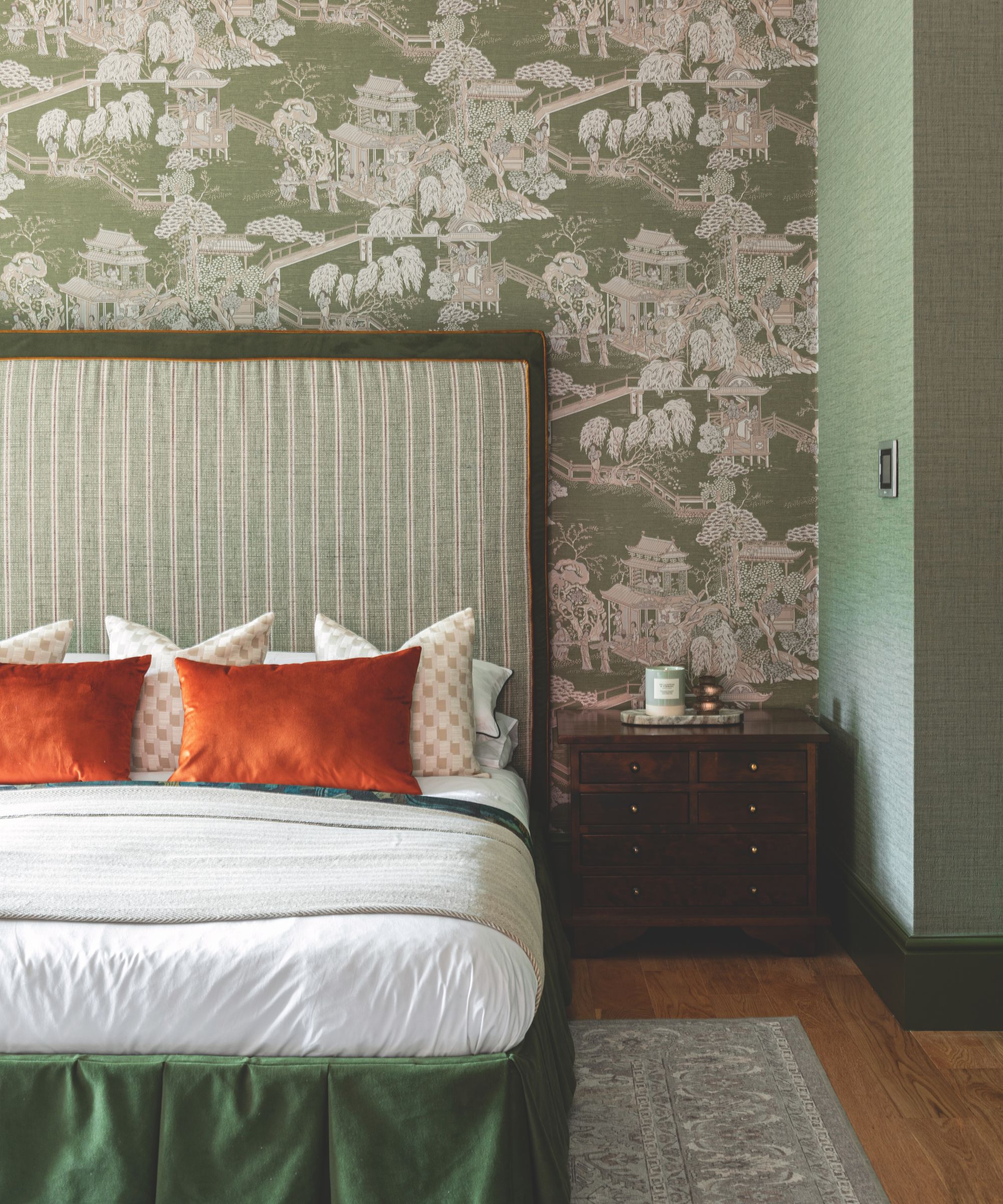 A maximalist bedroom scene featuring a tall, green striped headboard against Chinoiserie-style wallpaper. The bed is layered with crisp white linens and vibrant orange velvet pillows, set next to a dark wood bedside chest.