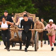 A group of people (l-r Eric Nam, Natalie Anderson, Michael Rapaport, Rob Rausch, Tara Lipinski, Candiace Dillard Bassett) push a throne-chariot with a man (Colton Underwood) sitting on it, in 'The Traitors' season 4.