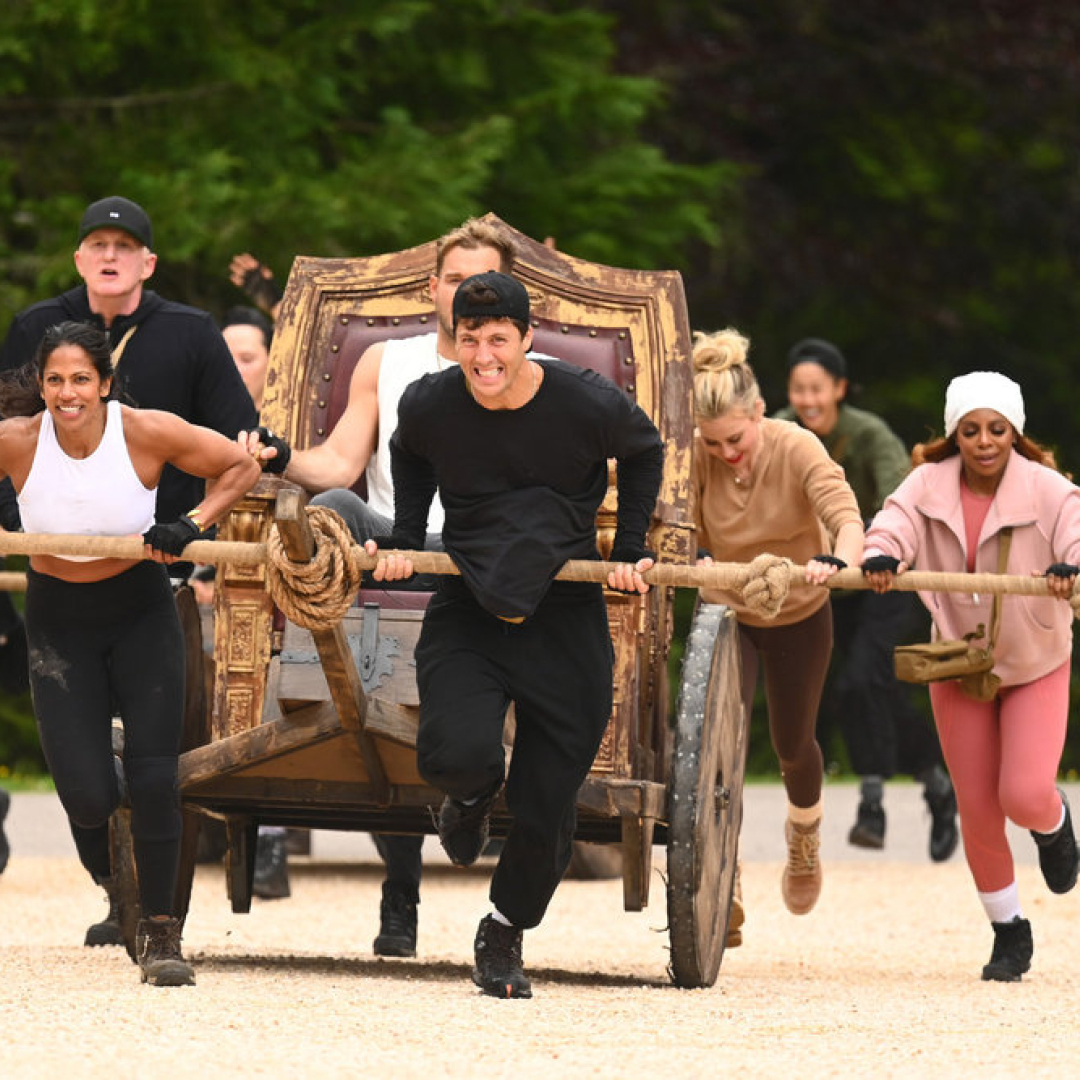 A group of people (l-r Eric Nam, Natalie Anderson, Michael Rapaport, Rob Rausch, Tara Lipinski, Candiace Dillard Bassett) push a throne-chariot with a man (Colton Underwood) sitting on it, in 'The Traitors' season 4.