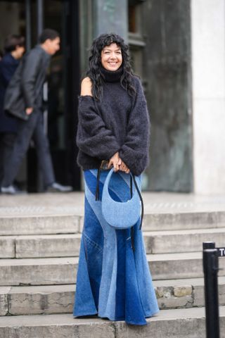 A guest wears a black turtleneck fluffy / cut-out shoulder details pullover, a dark and pale blue embroidered bicolored large fringed midi skirt, green fluffy platform soles / high heels ankle shoes, a pale blue denim handbag from Coperni , outside Awake, during Paris Fashion Week - Womenswear Fall Winter 2023 2024, on March 07, 2023 in Paris, France.