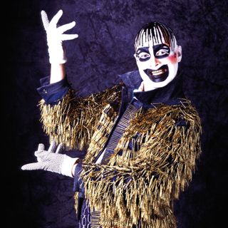 Leigh Bowery wearing black and purple and white face makeup and a jacket covered in gold tinsel