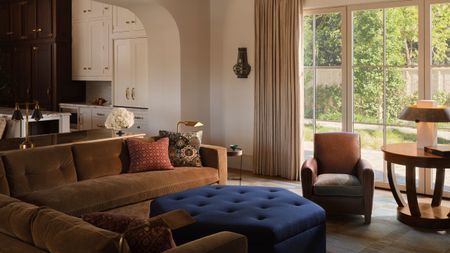 an open plan family room and kitchen with doors onto the garden, anchored by a toffee velvet L-shaped couch with blue hexagonal ottoman and leather chair