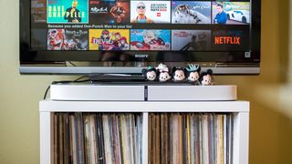 En vit Sonos Playbase ligger på en liten vit TV-bänk med en TV på väggen ovanför.