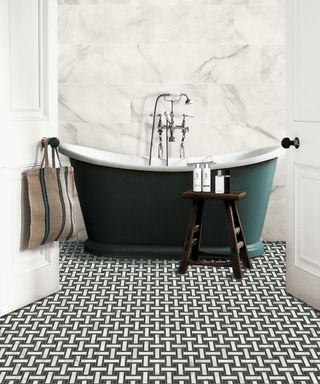 A dark green free standing bath in front of marble wall tiles and small black and white mosaic floor tiles