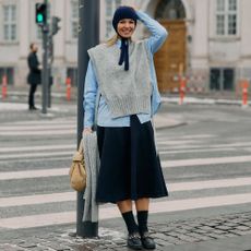 Woman poses while leaning on street lamp in front of a cross walk. She wears black skirt, button down shirt, gray sweater vest, and knitted babushka hat. 