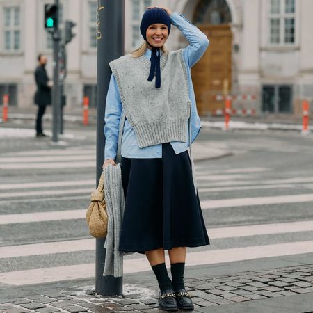 Woman poses while leaning on street lamp in front of a cross walk. She wears black skirt, button down shirt, gray sweater vest, and knitted babushka hat. 