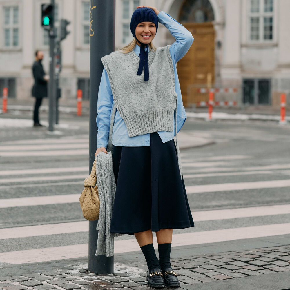 Woman poses while leaning on street lamp in front of a cross walk. She wears black skirt, button down shirt, gray sweater vest, and knitted babushka hat. 