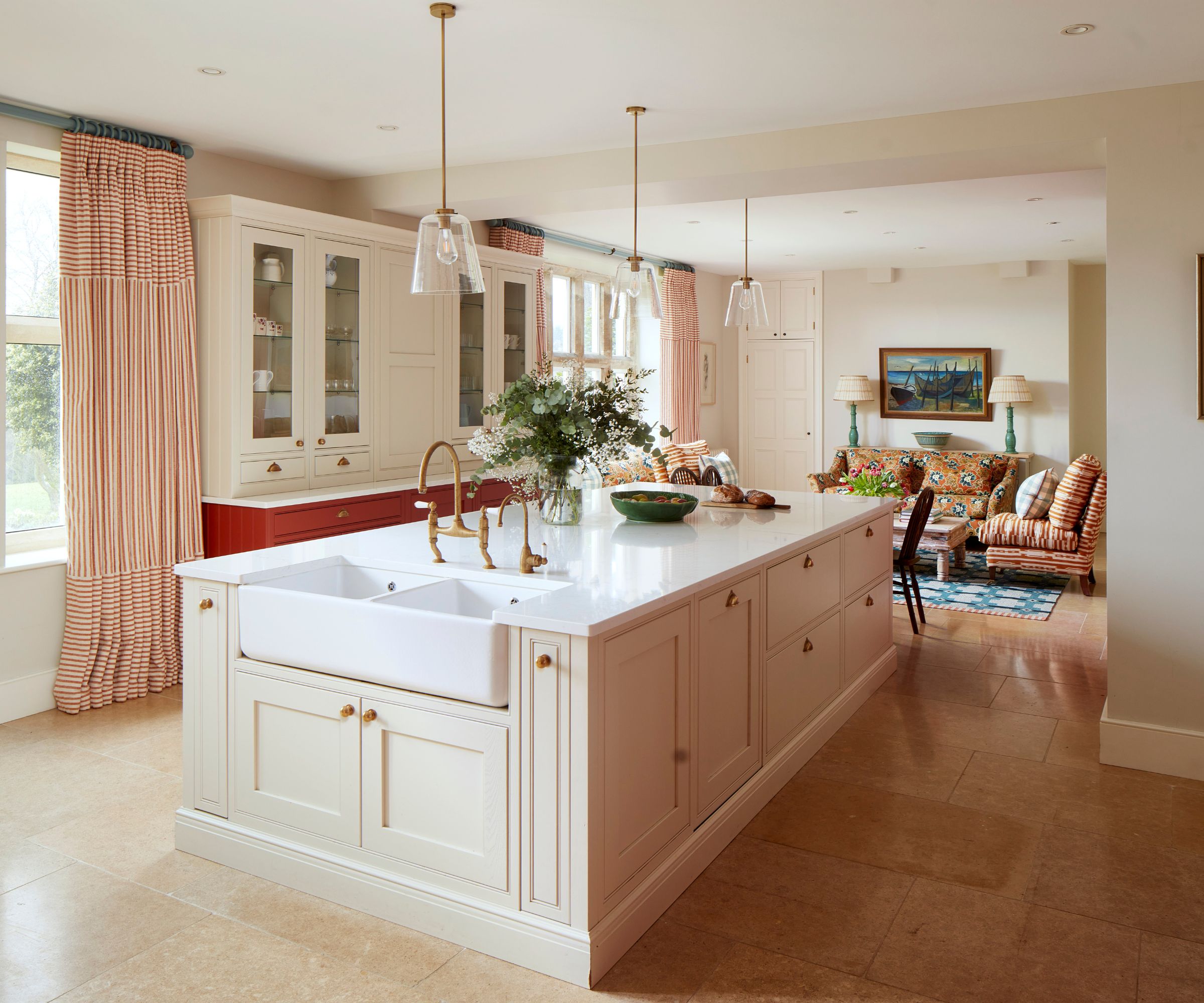 A soft kitchen with cream cabinetry, red and white striped curtains, glass-fronted cabinets, and a cozy seating area featuring a patterned sofa and armchair, a wooden coffee table, and two green table lamps