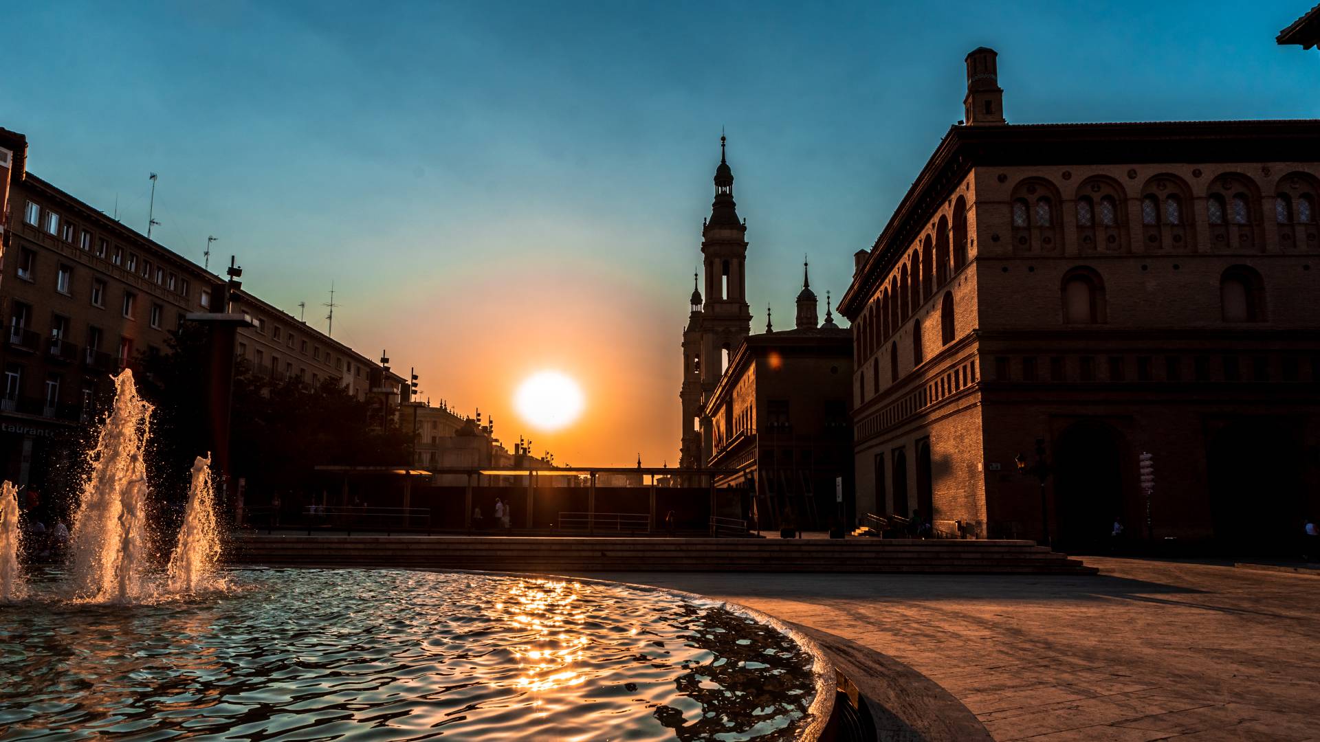 A city square with a fountain in the foreground and historic buildings silhouetted by a beautiful sunset in the background.