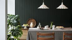 A dining table with a black and white check cloth over it. Wooden-backed chairs on either side of it. A monstera plant in the left corner, and two white milk glass pendants hanging above. 