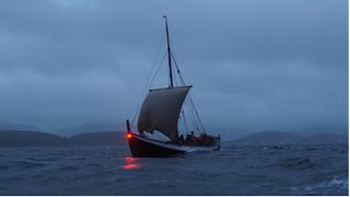 A recreation of a medieval era Norse ship sails on dark waters, with mountains in the background. 