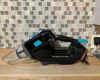 Charging the Bissell Pet Stain Eraser Duo portable carpet cleaner on wooden kitchen worktop with tiled kitchen wall in background