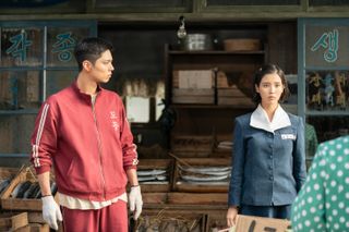 A man in a tracksuit (Park Bo-gum) and a woman in a school uniform (IU) stand in front of a rustic Korean show, in 'When Life Gives You Tangerines.'