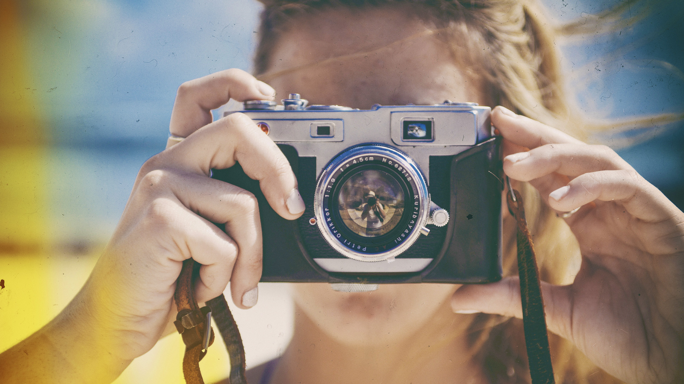 A close-up of a person&#039;s hands holding a vintage camera, capturing a sunny beach scene with a blurred colorful background