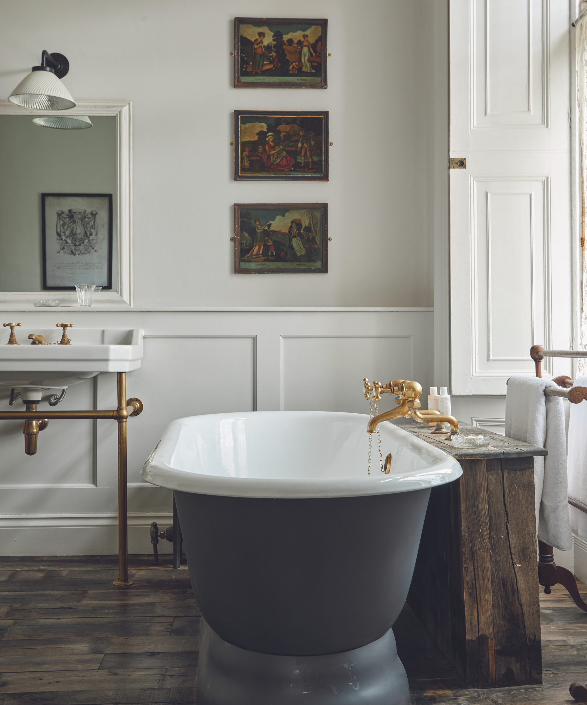 bathroom with white walls with panelling and artworks on wall and sink in background with mirror and wall lights above and and grey freestanding bath