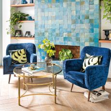 Living room with turquoise tiled chimney breast with wooden shelves either side and two blue armchairs beside a glass coffee table