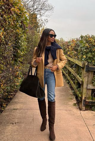 best suede boots shown on a photo of a woman standing in a park with turning leaves wearing a tan blazer over a tan t-shirt with a navy cardigan tied over the shoulders with skinny jeans tucked into dark brown suede knee-high boots and a suede handbag