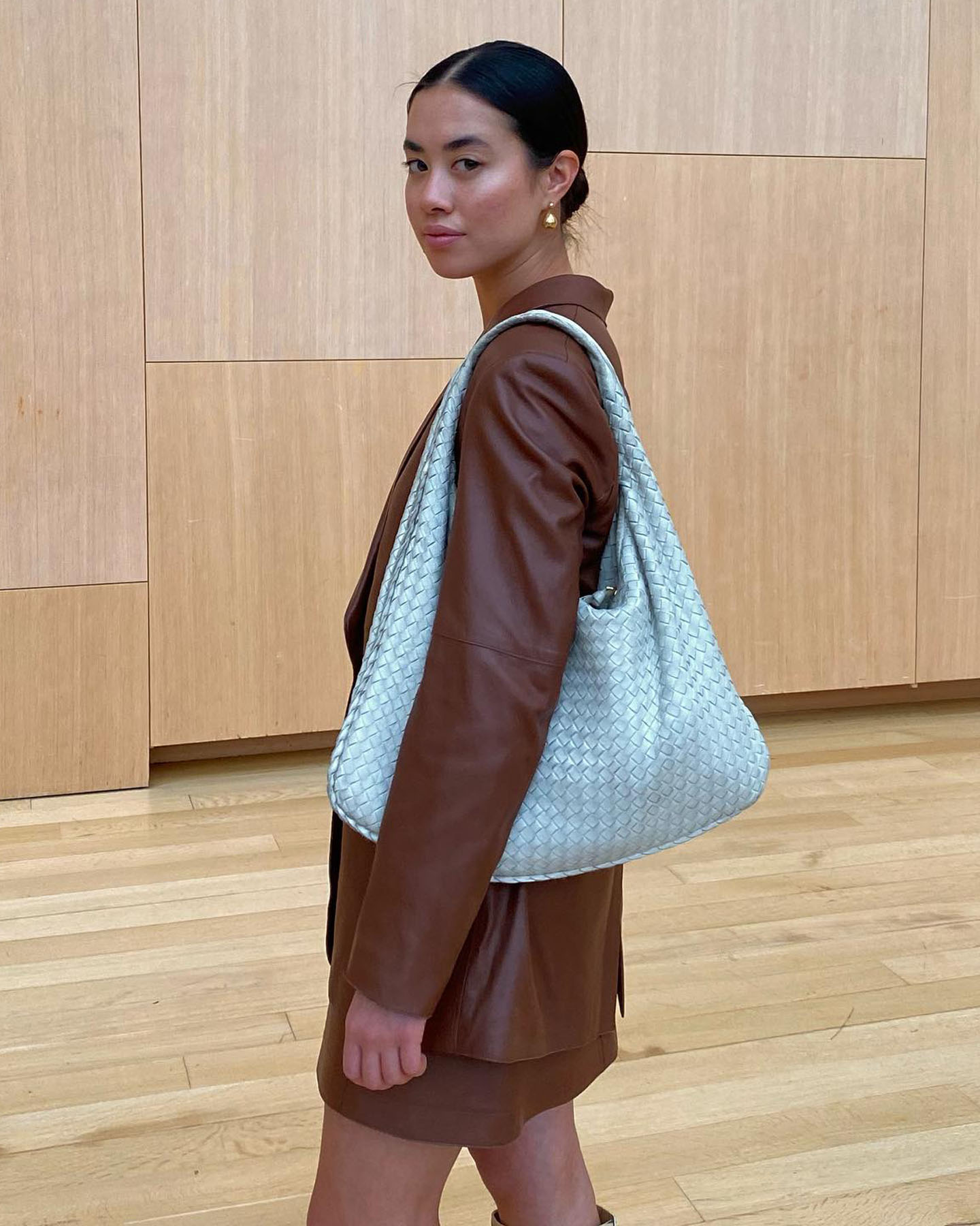 Influencer Sasha Mei poses in an all-wood room wearing a low bun, earrings, light blue Bottega Veneta Intrecciato Hobo Bag, and brown skirt suit.