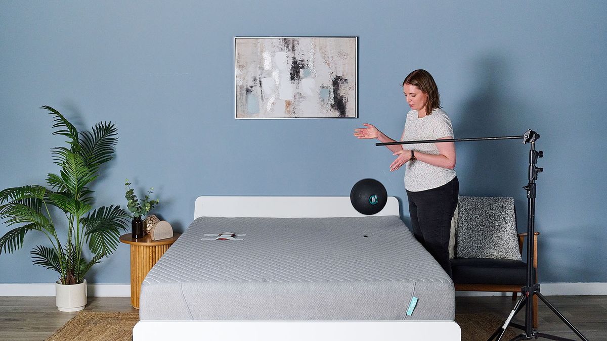 This image shows the objective slam ball motion isolation test in our sleep studio. The Siena Memory Foam Mattress on a bed frame sits in the center of a decorated room. To the right of the bed, our sleep writer Ruth Jones drops a 5kg slam ball onto the Siena Mattress. Next to her, a rig is set to mark 24 inches from the surface of the bed. On the left of the mattress is a mark roughly where a body would be when sleeping &mdash; this is the drop zone. On the left of the mattress, in the equivalent sleeping area, a phone with an accelerometer app open is taped to the bed.