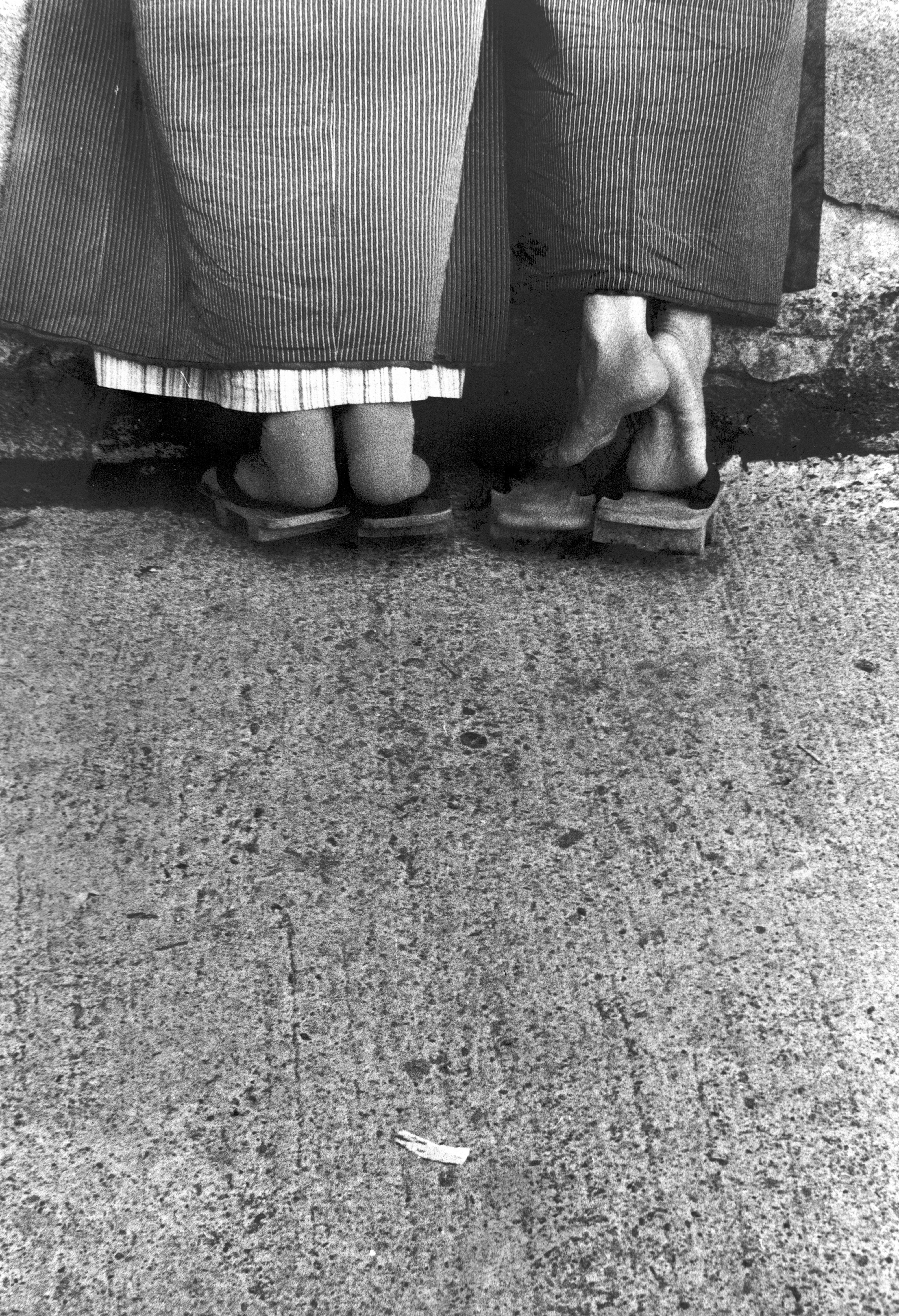 Black and white photograph of two pairs of feet in sandals, one standing on their tip toes.