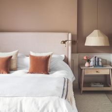 Terracotta painted bedroom with a pink bed frame, white bedding, and decorative cushions. There's a wooden bedside table with a hanging lampshade above it to the right of the bed