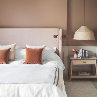 Terracotta painted bedroom with a pink bed frame, white bedding, and decorative cushions. There's a wooden bedside table with a hanging lampshade above it to the right of the bed