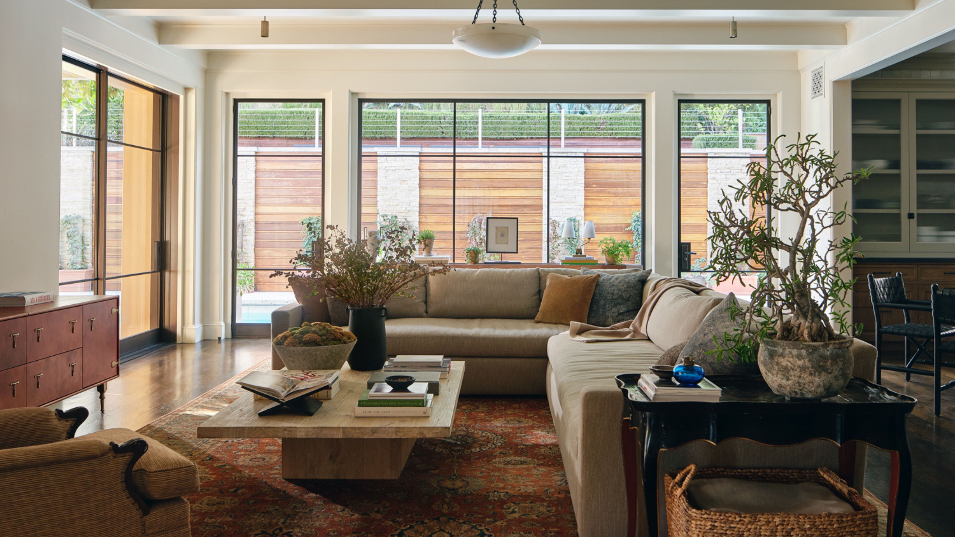a neutral family room with large glass windows and a sectional couch styled on a red vintage rug with a stone coffee table and layered decor