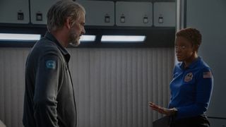 Woman wearing a blue jumpsuit is explaining something to a man (gray hair and beard) wearing a gray jacket.